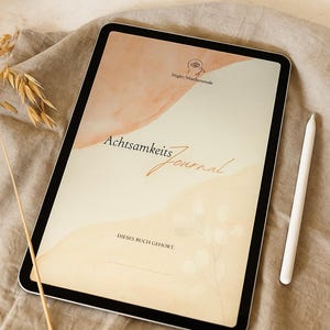May include: A digital tablet displaying a journal cover with the text "Achtsamkeits Journal" in gold script. The cover features a watercolor design in peach and cream tones. A white stylus rests beside the tablet.