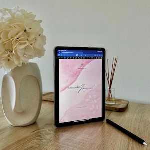 May include: A digital tablet displaying a pink and white journal cover with the text "Achtsamkeits Journal" and a stylus. A white floral arrangement sits in a decorative vase, and a reed diffuser is on a wooden surface.