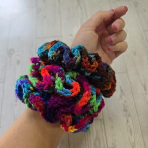 May include: A colorful, handmade crochet scrunchie. The scrunchie features a vibrant mix of rainbow colors including blue, green, purple, orange, and brown. The accessory is designed to hold hair in a ponytail or bun.