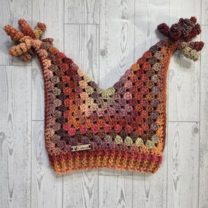 May include: A handmade crochet hat in a granny square pattern, featuring a mix of warm colors like orange, red, and brown. The hat has two decorative, textured elements on each side. A small tag with the word "Curious" is attached.