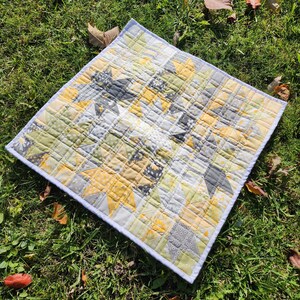 May include: A quilt with a patchwork design featuring yellow, gray, and white stars on a white background. The quilt is edged with a light blue trim.