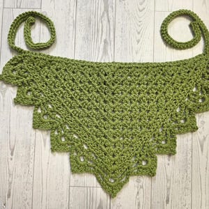May include: A handmade, green crochet top with a scalloped edge and two looped straps. The top is made of a textured yarn and is displayed on a light wood-grain surface. The crochet work features a detailed, open stitch pattern.