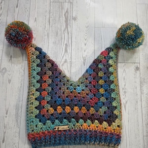 May include: A multi-colored crochet hat featuring a granny square pattern with two large yarn pom-poms at the top corners. The hat has a ribbed brim and a small rectangular wooden tag with the text Curious attached to the front lower edge.