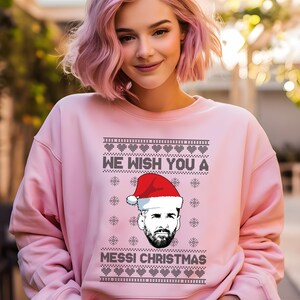 Puede incluir: Sudadera rosa claro con el texto negro "WE WISH YOU A MESSI CHRISTMAS". El dise&ntilde;o incluye un gr&aacute;fico de una persona con un gorro de Pap&aacute; Noel. La sudadera es de color rosa liso.