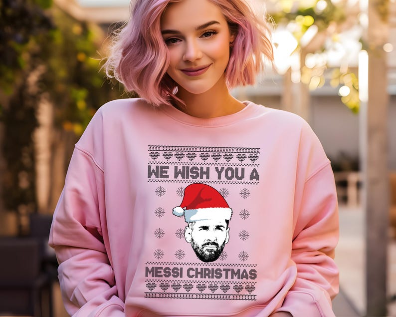 Puede incluir: Sudadera rosa claro con el texto negro "WE WISH YOU A MESSI CHRISTMAS". El dise&ntilde;o incluye un gr&aacute;fico de una persona con un gorro de Pap&aacute; Noel. La sudadera es de color rosa liso.