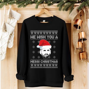 Puede incluir: Sudadera negra con el texto blanco "WE WISH YOU A MESSI CHRISTMAS". El dise&ntilde;o incluye un retrato de un hombre con gorro de Pap&aacute; Noel, copos de nieve y un patr&oacute;n festivo. La sudadera est&aacute; colgada en una percha de madera.