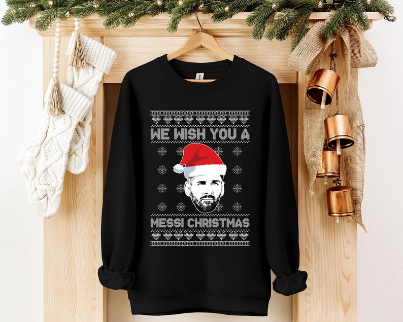 Puede incluir: Sudadera negra con el texto blanco "WE WISH YOU A MESSI CHRISTMAS". El dise&ntilde;o incluye un retrato de un hombre con gorro de Pap&aacute; Noel, copos de nieve y un patr&oacute;n festivo. La sudadera est&aacute; colgada en una percha de madera.