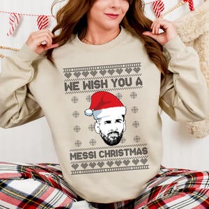 Puede incluir: Sudadera beige de cuello redondo con un dise&ntilde;o festivo. La sudadera presenta el texto "WE WISH YOU A MESSI CHRISTMAS" con un gr&aacute;fico de un hombre con un gorro de Pap&aacute; Noel. El dise&ntilde;o incluye corazones y copos de nieve.