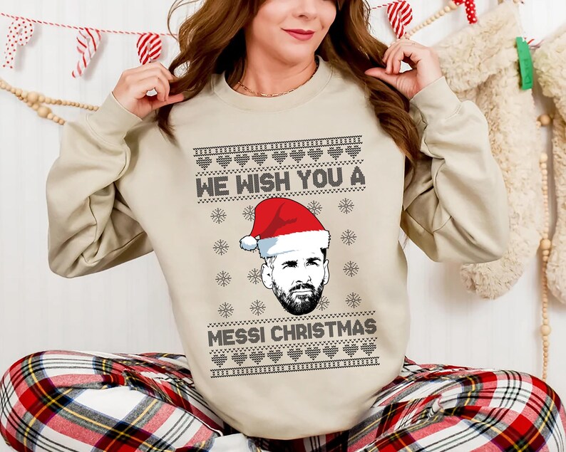Puede incluir: Sudadera beige de cuello redondo con un dise&ntilde;o festivo. La sudadera presenta el texto "WE WISH YOU A MESSI CHRISTMAS" con un gr&aacute;fico de un hombre con un gorro de Pap&aacute; Noel. El dise&ntilde;o incluye corazones y copos de nieve.
