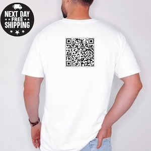 May include: White t-shirt with a black QR code printed on the back. A circular graphic with the text "NEXT DAY FREE SHIPPING" is on the upper left. The shirt is plain and has short sleeves.