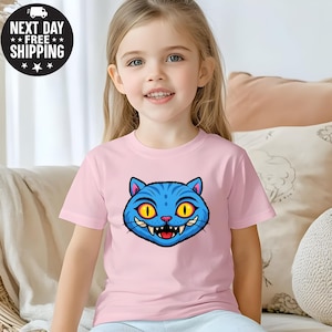May include: A light pink t-shirt featuring a cartoon blue cat face with yellow eyes and sharp teeth. The cat graphic is centered on the shirt. The shirt has short sleeves and a round neckline. The image also includes the text "NEXT DAY FREE SHIPPING".