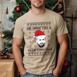 Puede incluir: Camiseta beige con un dise&ntilde;o festivo. La camiseta presenta el texto "WE WISH YOU A MESSI CHRISTMAS" sobre un gr&aacute;fico de una persona con un gorro de Pap&aacute; Noel. El dise&ntilde;o incluye copos de nieve y formas de coraz&oacute;n, creando un tema navide&ntilde;o.