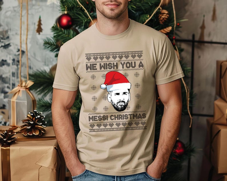 Puede incluir: Camiseta beige con un dise&ntilde;o festivo. La camiseta presenta el texto "WE WISH YOU A MESSI CHRISTMAS" sobre un gr&aacute;fico de una persona con un gorro de Pap&aacute; Noel. El dise&ntilde;o incluye copos de nieve y formas de coraz&oacute;n, creando un tema navide&ntilde;o.