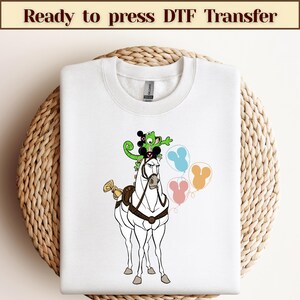 Könnte beinhalten: Weißes T-Shirt mit einer Cartoon-Abbildung eines Pferdes, eines Chamäleons und Mickey Mouse. Das Pferd trägt einen Sattel und ein Zaumzeug. Das Chamäleon sitzt auf Mickey Mouse's Kopf. Im Hintergrund befinden sich drei Ballons.