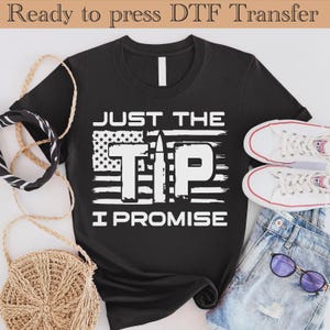 May include: Black t-shirt with the text "JUST THE TIP I PROMISE" in white, featuring an American flag design and a bullet graphic. The shirt is paired with denim shorts, white sneakers, and sunglasses.