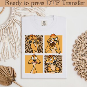 Op de afbeelding: Wit T-shirt met een ontwerp van vier panelen met een cartoon stokstaartje in verschillende poses. Het stokstaartje is oranje en bruin met een rode haarlok. De panelen wisselen af tussen een luipaardprint achtergrond en een effen oranje achtergrond. De tekst "Ready to press DTF Transfer" staat bovenaan.