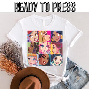 以下が含まれることがあります： 漫画のプリンセスの顔がカラフルに描かれた白いTシャツ。 シャツは袖をまくり上げており、上部には「READY TO PRESS」の文字。 茶色の帽子とデニムショーツも画像に写っています。