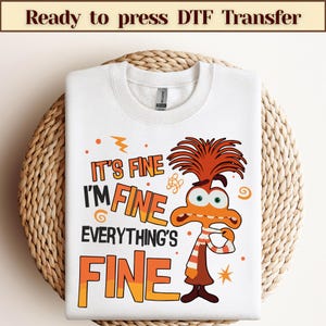 以下が含まれることがあります： 「IT'S FINE I'M FINE EVERYTHING'S FINE」の文字と、オレンジ色の髪とストライプのスカーフをした漫画のキャラクターが描かれた白いスウェットシャツ。スウェットシャツは、織られた円形の表面に表示されています。