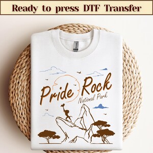 Może przedstawiać: Biała koszulka z nadrukiem graficznym przedstawiającym lwa na górze z tekstem "Pride Rock National Park".