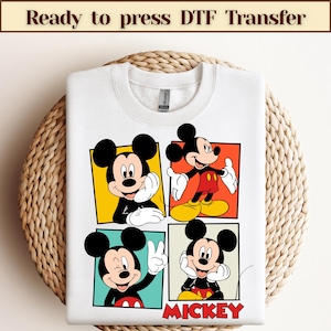 May include: White sweatshirt featuring a colorful Mickey Mouse design. The graphic includes four panels with different poses of Mickey Mouse. The word "MICKEY" is printed in red at the bottom. The text "Ready to press DTF Transfer" is at the top.