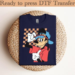 Puede incluir: Camiseta azul marino con un personaje de dibujos animados con una sudadera roja y una gorra azul, sobre un fondo a cuadros. El texto "Ready to press DTF Transfer" está en la parte superior. La camiseta está doblada sobre una superficie tejida.