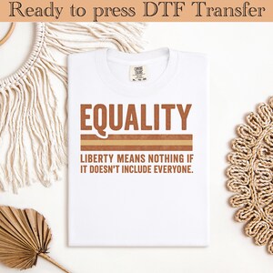 Puede incluir: Camiseta blanca con la palabra "EQUALITY" en letras marrones sobre una franja marrón y beige. Debajo, la camiseta dice "LIBERTY MEANS NOTHING IF IT DOESN'T INCLUDE EVERYONE."