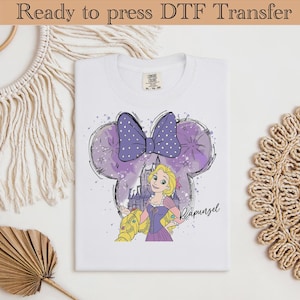 Op de afbeelding: Wit T-shirt met een paarse strik met witte stippen en een Rapunzel-afbeelding. Het ontwerp bevat een kasteel en de naam "Rapunzel" in script. Het shirt is gelabeld "Ready to press DTF Transfer".