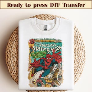 Może przedstawiać: Biała bluza z okrągłym dekoltem z okładką komiksu Spider-Man w stylu vintage. Grafika zawiera tekst "The Amazing Spider-Man" i "Marvel Comics Group". Bluza jest prezentowana na tkanej, okrągłej powierzchni.