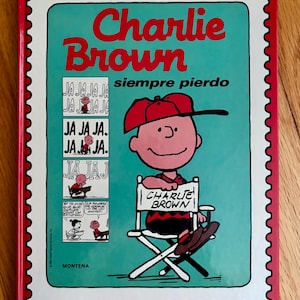 May include: A book cover featuring Charlie Brown, wearing a red cap, sitting in a director's chair. The title "Charlie Brown" is in red, with the Spanish phrase "siempre pierdo" below. The cover has a red border with a postage stamp design.