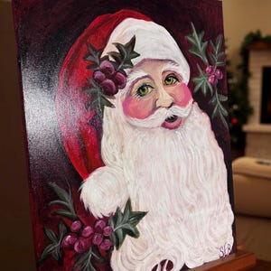 Może przedstawiać: Obraz Świętego Mikołaja z białą brodą i czapką, różowymi policzkami i zielonymi oczami. Dzieło ma czerwone tło i jest ozdobione gałązkami ostrokrzewu i jagodami. Obraz jest wystawiony na drewnianym sztaludze.