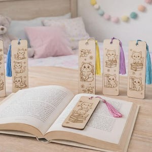 Puede incluir: Marcadores de libros de madera con ilustraciones de gatos grabadas. Cada marcador presenta un diseño de gato diferente, algunos con libros y flores, y borlas de colores. Los marcadores se muestran con un libro abierto sobre una superficie de madera.