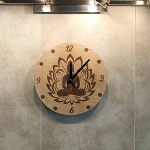 Può includere: Un orologio in legno con sfondo marrone e una silhouette nera di una figura di Buddha in meditazione circondata da un motivo a fiore di loto. Il quadrante dell'orologio ha lancette nere e numeri neri.