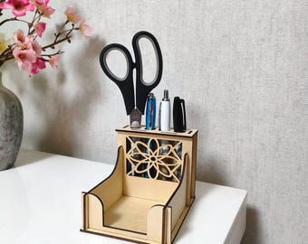 Puede incluir: Un organizador de escritorio de madera con un diseño floral cortado con láser. Contiene tijeras negras, bolígrafos y otros útiles de oficina. El organizador es de madera clara y está sobre una superficie blanca.