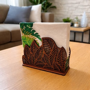 Può includere: Un portatovaglioli in legno marrone con un design a foglie traforate. Contiene tovaglioli bianchi con un motivo a foglie verdi e oro. Il portatovaglioli è su un tavolo di legno marrone chiaro.