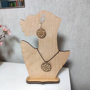 May include: A wooden jewelry display featuring a necklace and earrings. The necklace has a circular pendant with a detailed floral design. The earrings mirror the pendant design. The display is a light wood color, shaped like a bust.