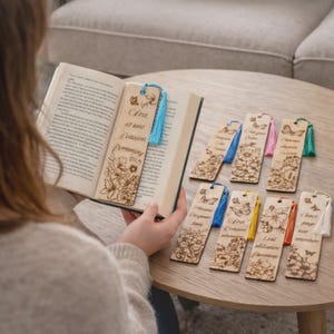 Peut inclure: Marque-pages en bois avec des motifs floraux et de papillons, certains avec du texte en français. Chaque marque-page a un pompon coloré. Un marque-page est placé dans un livre ouvert. Les marque-pages sont sur une table ronde en bois.