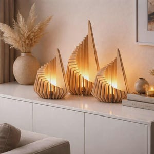 Può includere: Tre lampade in legno dal design geometrico. Le lampade sono realizzate in legno chiaro e hanno un bagliore caldo e invitante. Le lampade sono disposte su una superficie bianca, con un vaso di fiori secchi sullo sfondo.
