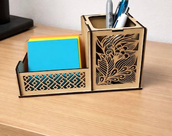 Puede incluir: Un organizador de escritorio de madera con dos compartimentos. Una sección contiene notas adhesivas de colores, y la otra tiene bolígrafos y lápices. El organizador presenta patrones decorativos recortados y diseños florales.