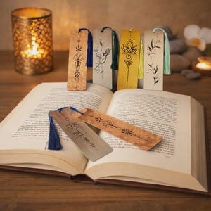 Peut inclure: Un livre ouvert avec plusieurs marque-pages décoratifs en métal présentant des motifs complexes et des pompons colorés. Les signets ont des motifs floraux et géométriques. Une bougie allumée dans un photophore est en arrière-plan.