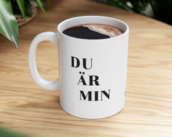 May include: White ceramic coffee mug with black text that reads "DU ÄR MIN". The mug is filled with coffee and is sitting on a wooden surface.