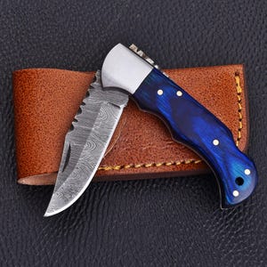 May include: A folding pocket knife with a Damascus steel blade featuring a distinct wavy pattern and a handle made of polished blue resin with gold-coloured rivets. The knife is positioned against a brown leather sheath with visible stitching.