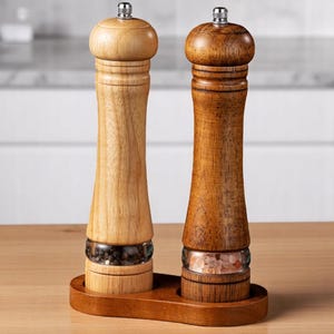 May include: A wooden salt and pepper grinder set. One grinder is light-colored, the other is dark-colored. Both have a glass section to view the contents and sit in a wooden tray. The salt grinder contains pink salt.