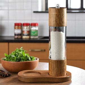 Op de afbeelding: Een houten zout- of pepermolen met een helder glazen midden gevuld met zout. De molen staat op een houten voet met een kleine cirkelvormige inkeping. Een houten kom met groene kruiden en een houten kom staan ook op de tafel.