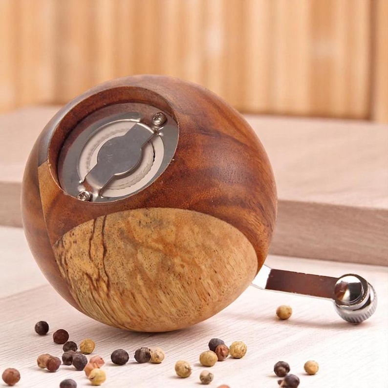 K&ouml;nnte beinhalten: Ein runder, brauner und hellbrauner Pfefferm&uuml;hle aus Holz mit einer silbernen Metallkurbel. Die M&uuml;hle steht auf einer wei&szlig;en Oberfl&auml;che mit verstreuten Pfefferk&ouml;rnern.