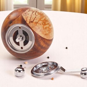 K&ouml;nnte beinhalten: Ein runder, brauner und hellbrauner Pfefferm&uuml;hle aus Holz. Die M&uuml;hle hat einen Metallmechanismus und einen silbernen Griff. Die M&uuml;hle ist zerlegt und die Teile befinden sich auf einer wei&szlig;en Oberfl&auml;che.