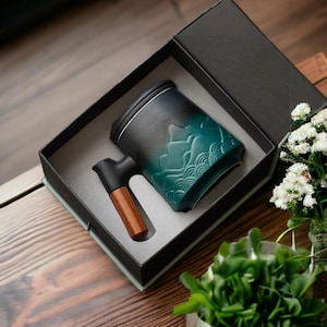 Puede incluir: Una taza de té de cerámica negra y verde con un asa de madera, en una caja de regalo negra. La taza de té tiene un diseño decorativo de montañas y olas.