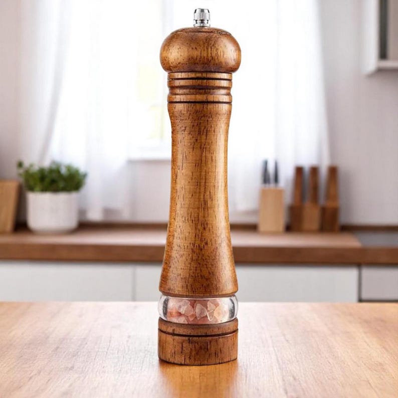 May include: A wooden salt or pepper grinder with a classic design. The grinder is made of light brown wood with a silver top and a clear glass section showing pink salt crystals. The grinder is on a wooden surface.
