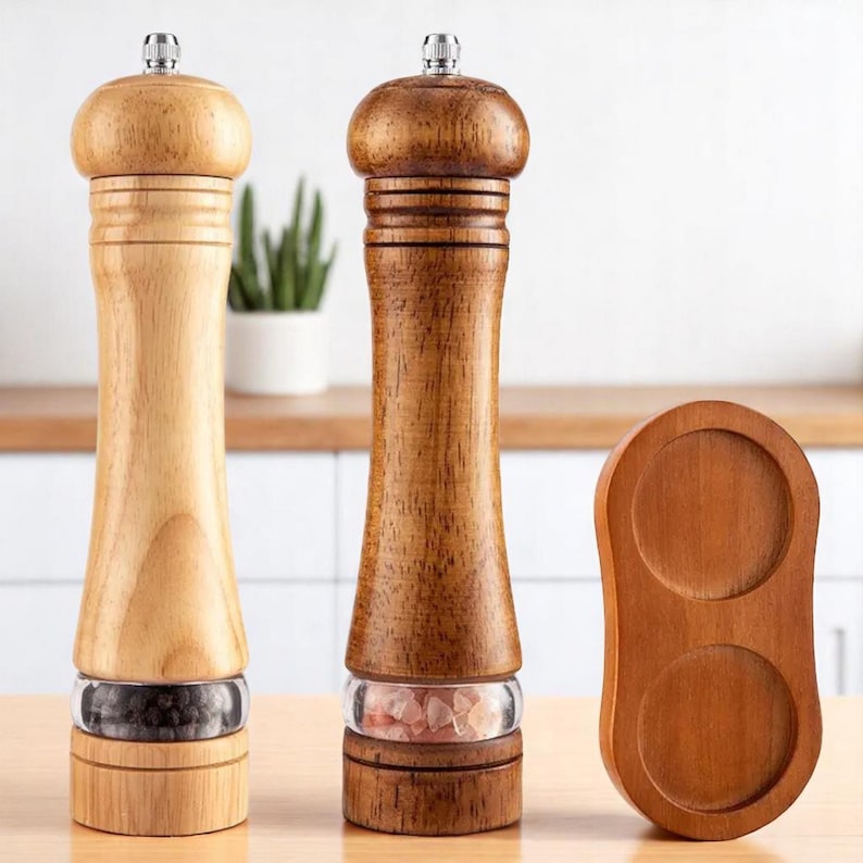 May include: Two wooden salt and pepper grinders with a clear glass window to view the contents. One is light-colored, the other is dark. A wooden holder with two circular indentations sits to the side. The grinders are topped with a silver metal cap.