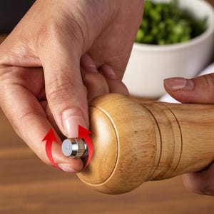 May include: A wooden pepper mill with a silver-colored adjustment knob. The mill is being adjusted by a person's hand. The wood has a light brown color and a smooth finish. The background includes a white bowl and green herbs.