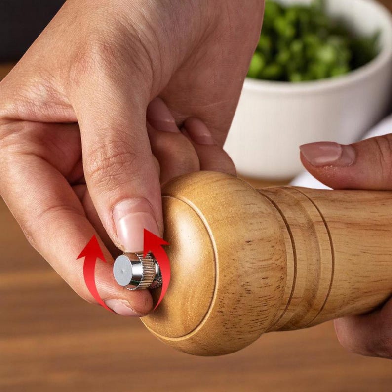 May include: A wooden pepper mill with a silver-colored adjustment knob. The mill is being adjusted by a person's hand. The wood has a light brown color and a smooth finish. The background includes a white bowl and green herbs.